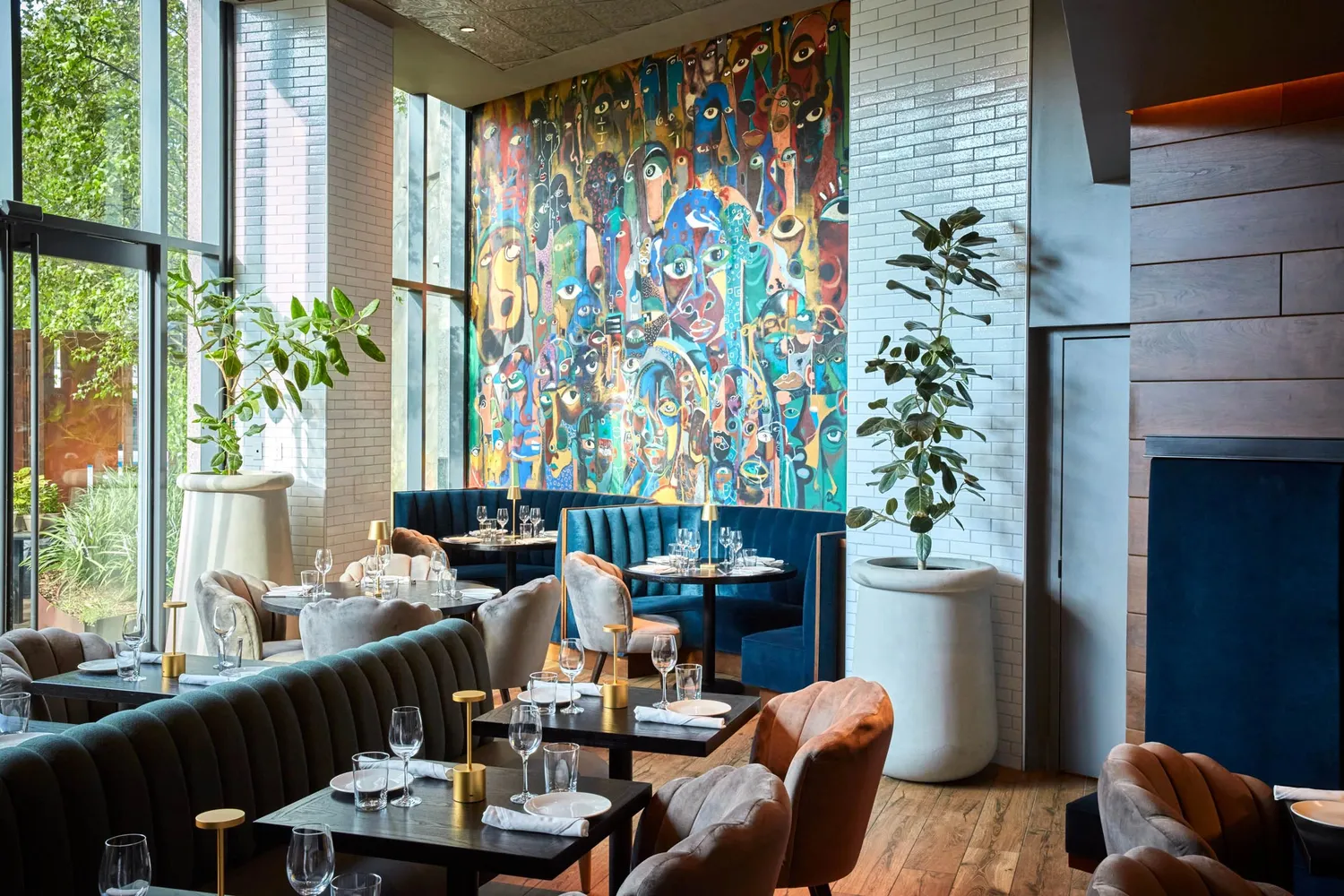 A modern restaurant interior with large windows, vibrant mural, and elegant table settings featuring dark velvet seating and tall plants in white pots. Upscale Caribbean Restaurant